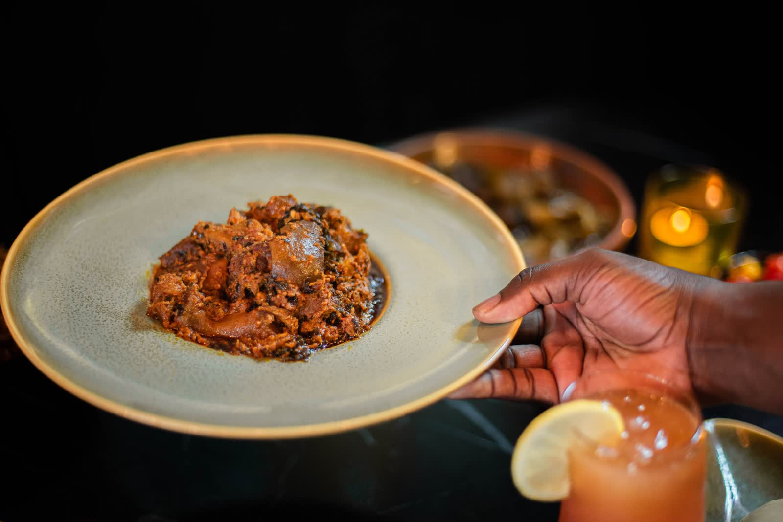 A hand presenting a plate of dark stew, a cocktail in the foreground
