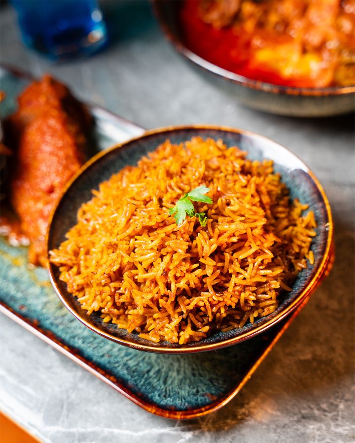 A bowl of jollof rice, with stew and a roll behind it