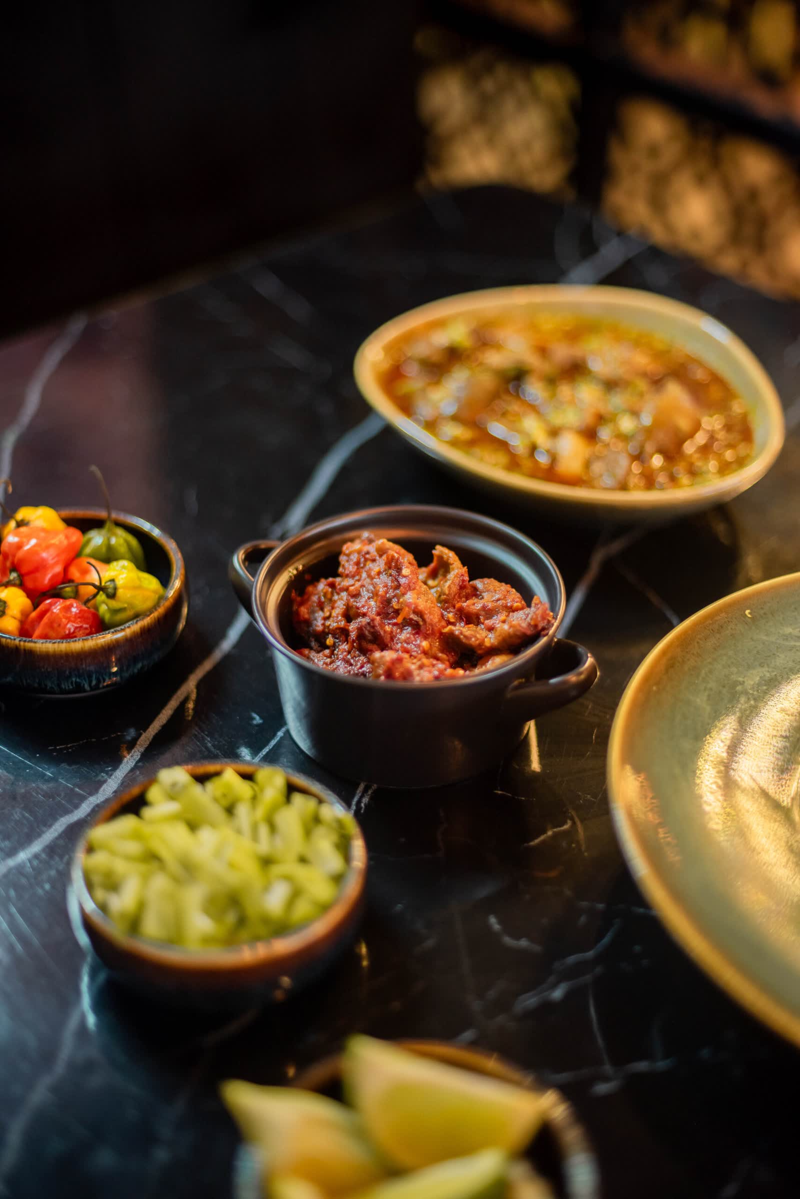 A spread on black marble — a bowl of stew with okra, small dishes of coloured peppers, chopped greens and lime wedges