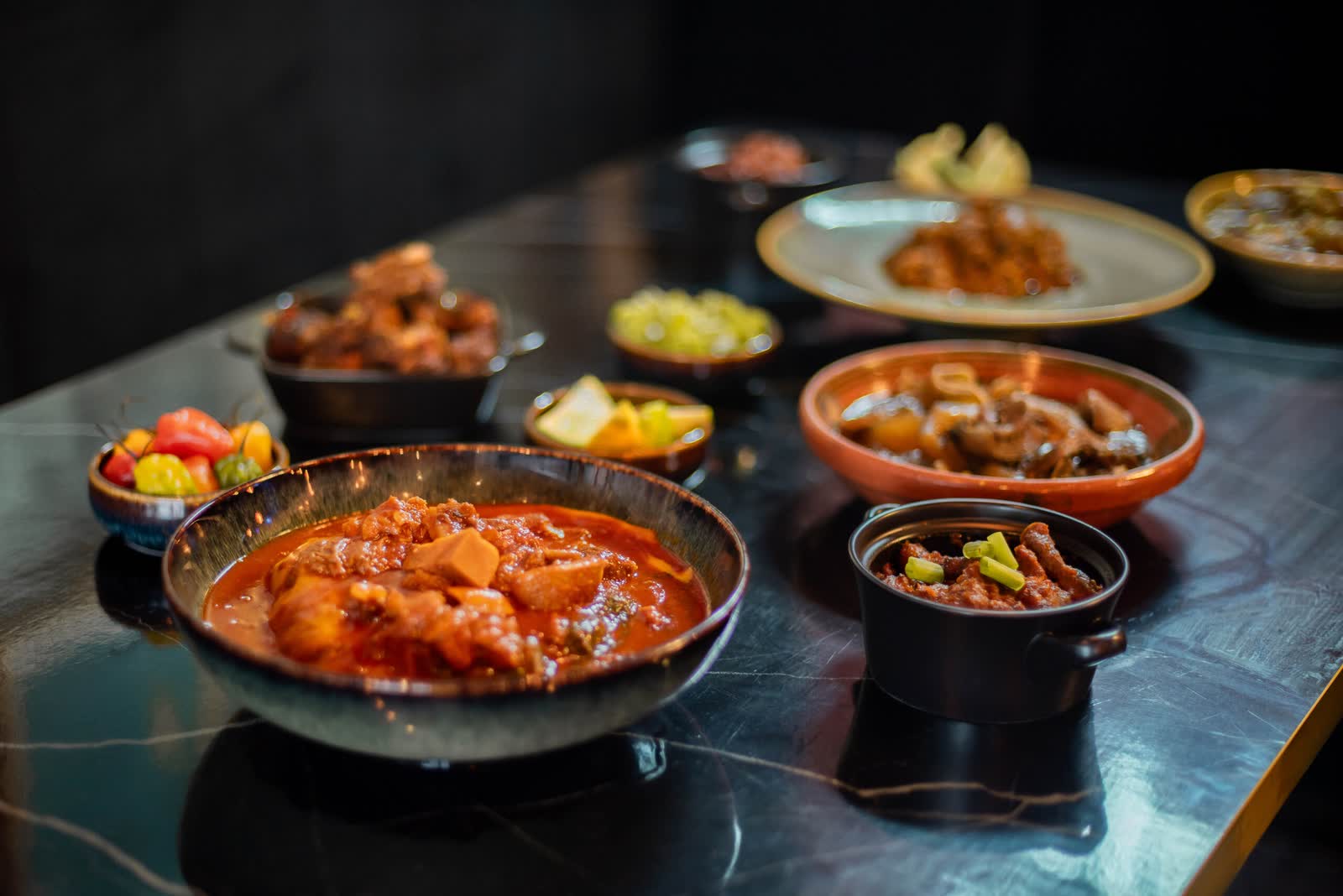 A shallow pan of red stew with chicken on black marble, ringed by small dishes