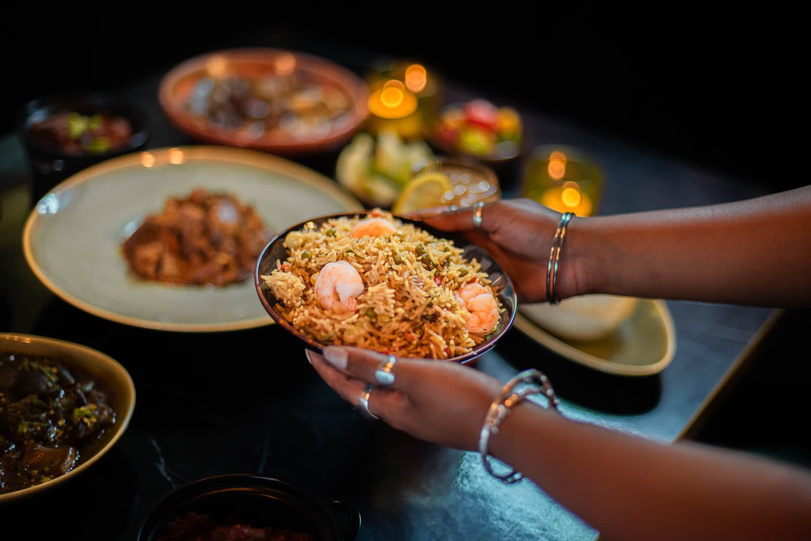Two hands, ringed and braceleted, holding out a bowl of rice topped with a prawn