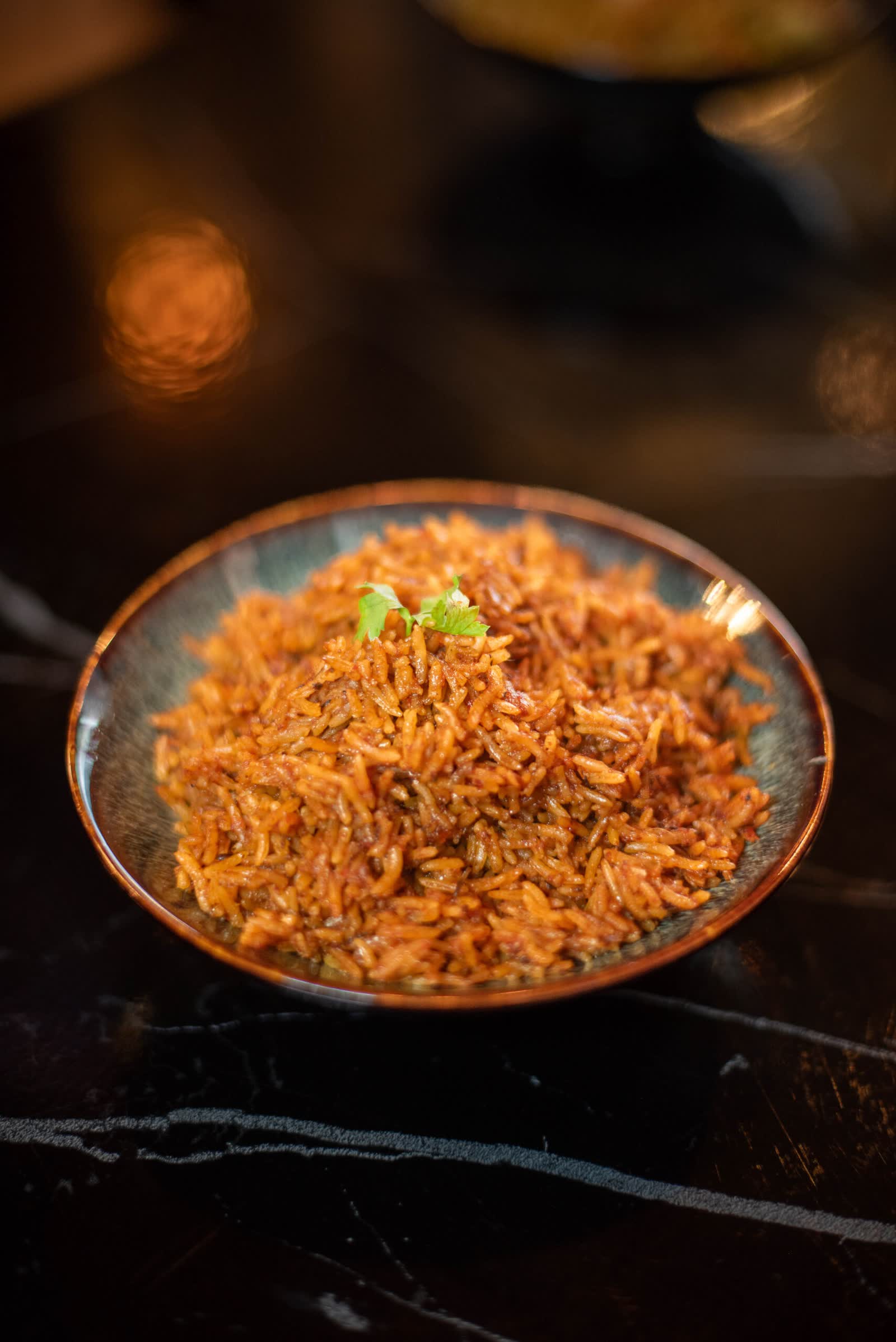 A bowl of orange-red rice with a herb sprig, on a dark plate