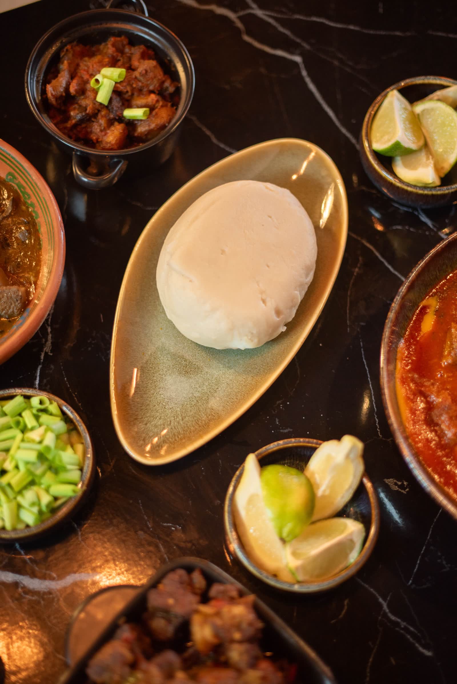 A smooth round of pounded dough on an oval plate, bowls of stew, greens and lime around it