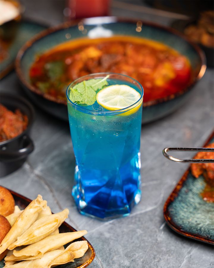 A tall blue cocktail with lime, set beside a bowl of stew